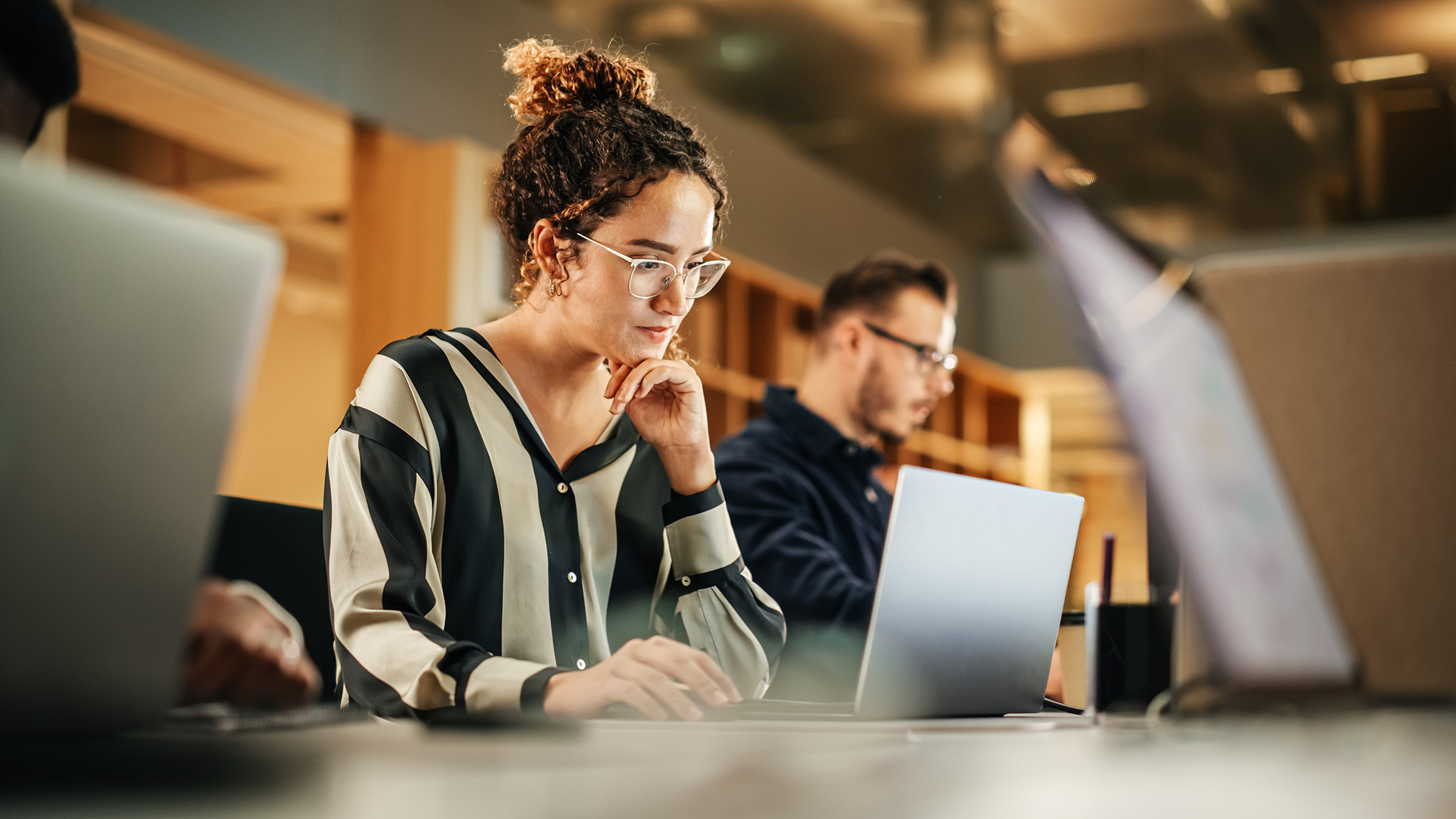 Enisca Browne - Woman wearing glasses works on a laptop in a modern office, with colleagues in the background.