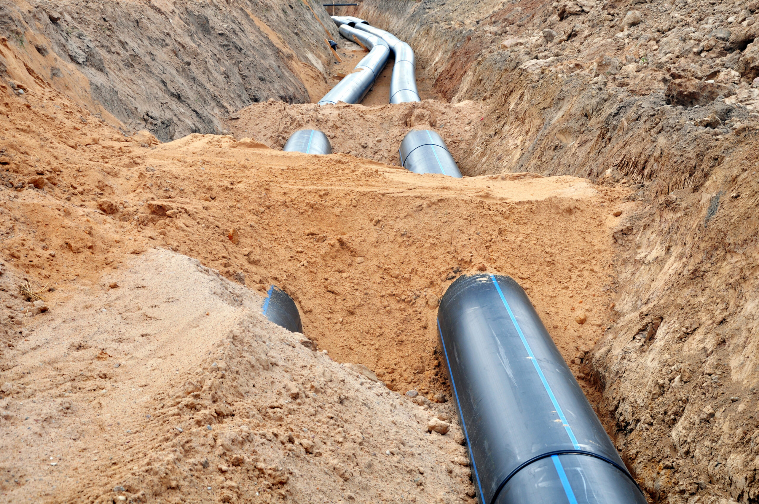 Enisca Browne - Large black pipes being installed underground in a trench through dirt and rocky soil.