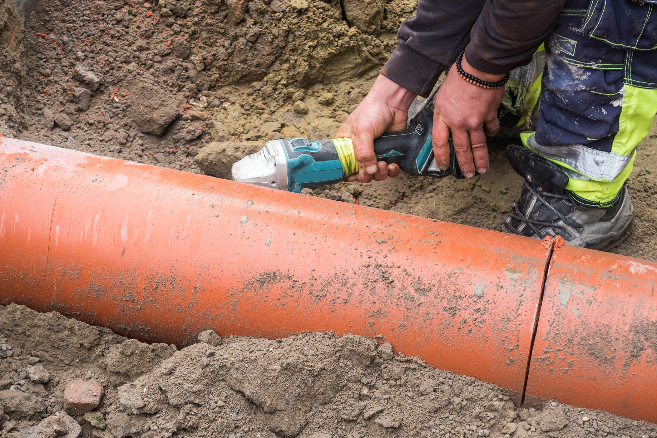 Enisca Browne - Worker using a power tool to cut an orange plastic pipe in a dirt trench.