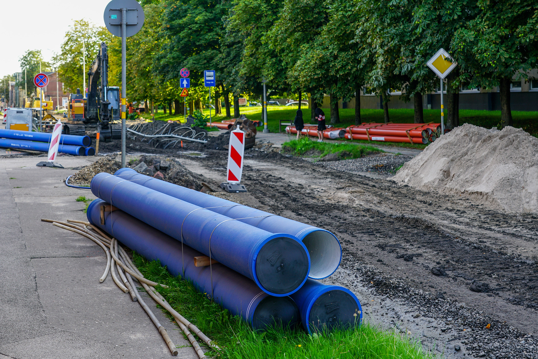 Enisca Browne - Construction site with large blue pipes, road barriers, and piles of dirt along a partially dug-up street.
