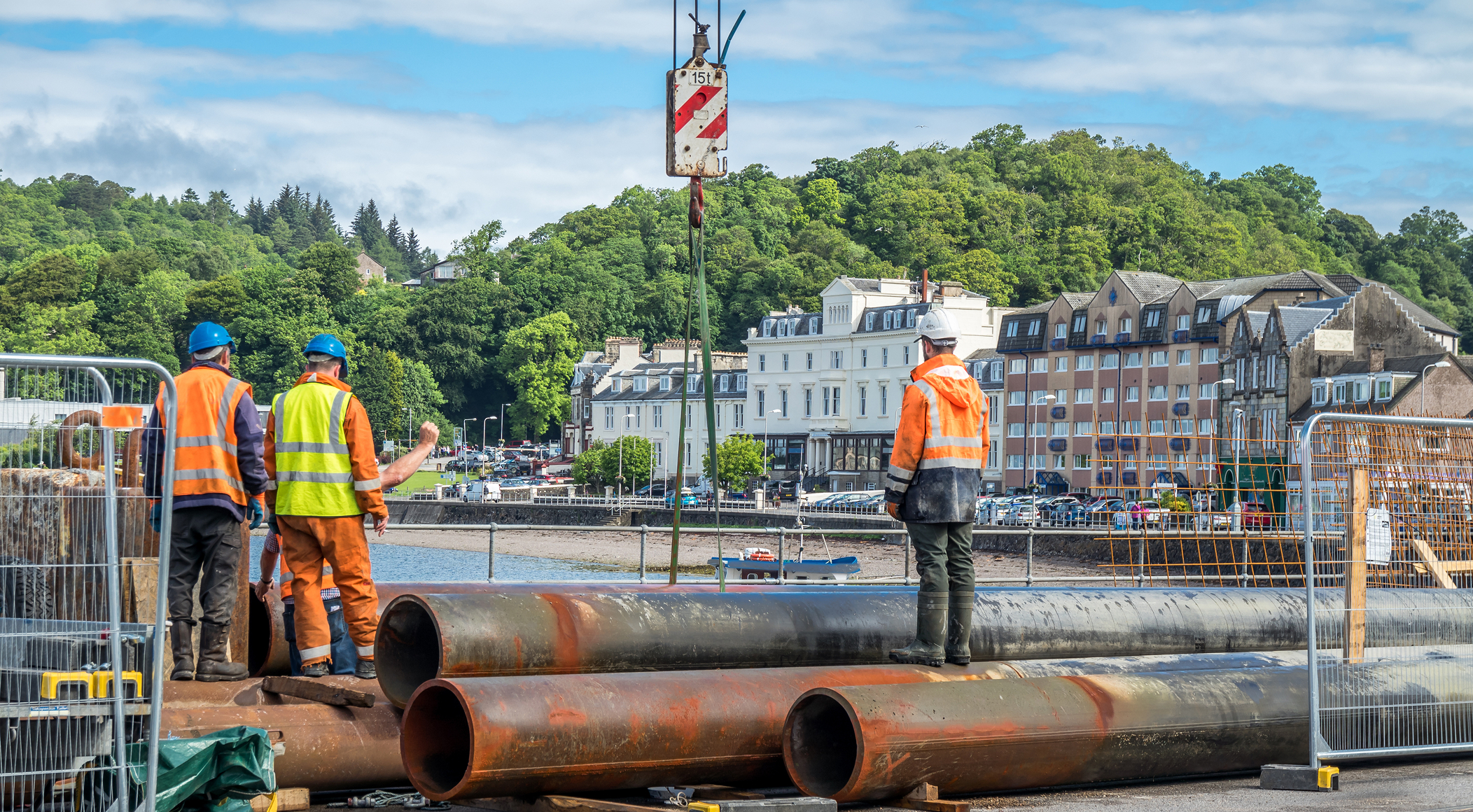 Enisca Browne - Construction workers in safety gear stand near large pipes at a waterfront site, with buildings in the background.