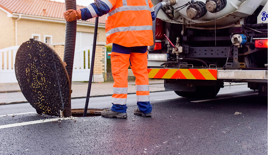 Enisca Browne - Worker in orange safety gear cleans a street sewer with a large hose from a specialized truck.