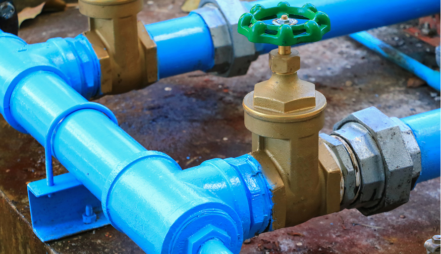 Enisca Browne - Close-up of blue water pipes with a brass valve and a green handle, set on a concrete surface.