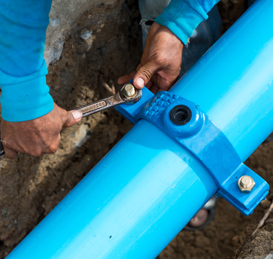 Enisca Browne - Person tightening bolts on a large blue water pipe with a wrench, outdoors on a construction site.