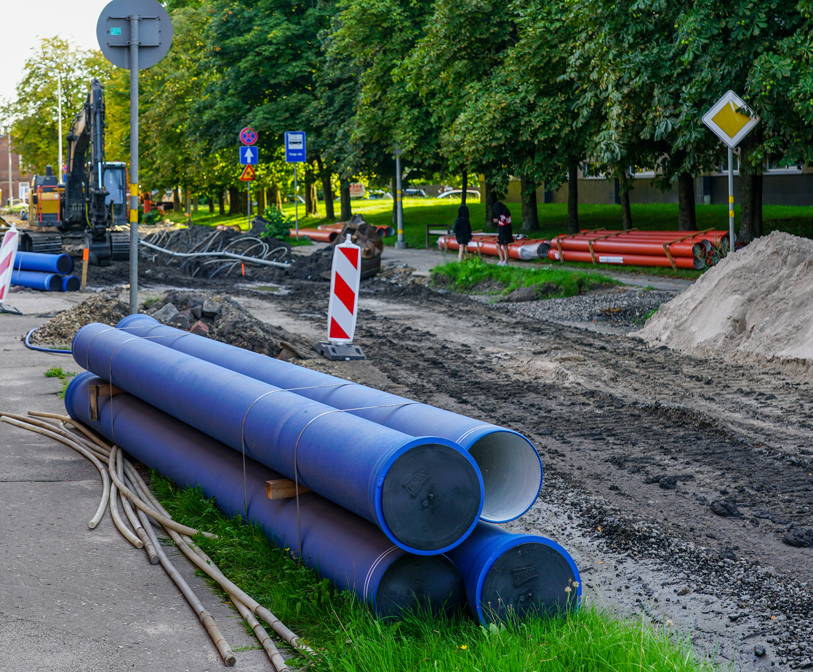Enisca Browne - Large blue pipes stacked beside a road under construction with dirt piles and construction equipment nearby.