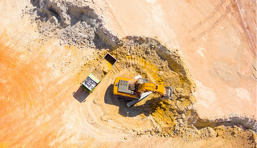 Enisca Browne - Aerial view of an excavator loading dirt into a dump truck at a construction or mining site.