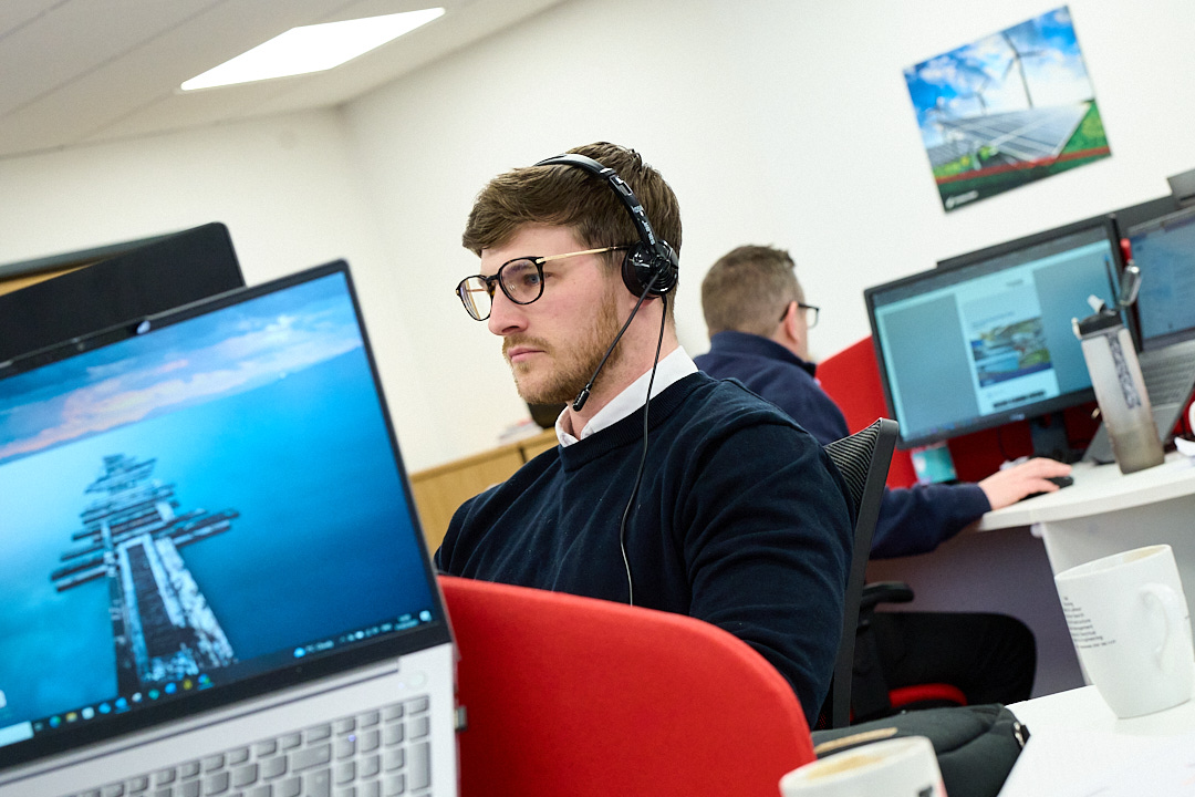 Enisca Browne - Man wearing glasses and headphones working at a computer in a modern office with colleagues nearby.