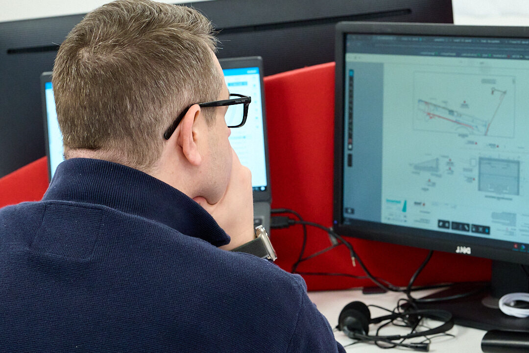 Enisca Browne - A person with glasses works at a desk, viewing diagrams on dual computer monitors.