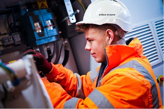 Enisca Browne - Man in orange safety jacket and white helmet working on electrical equipment indoors.