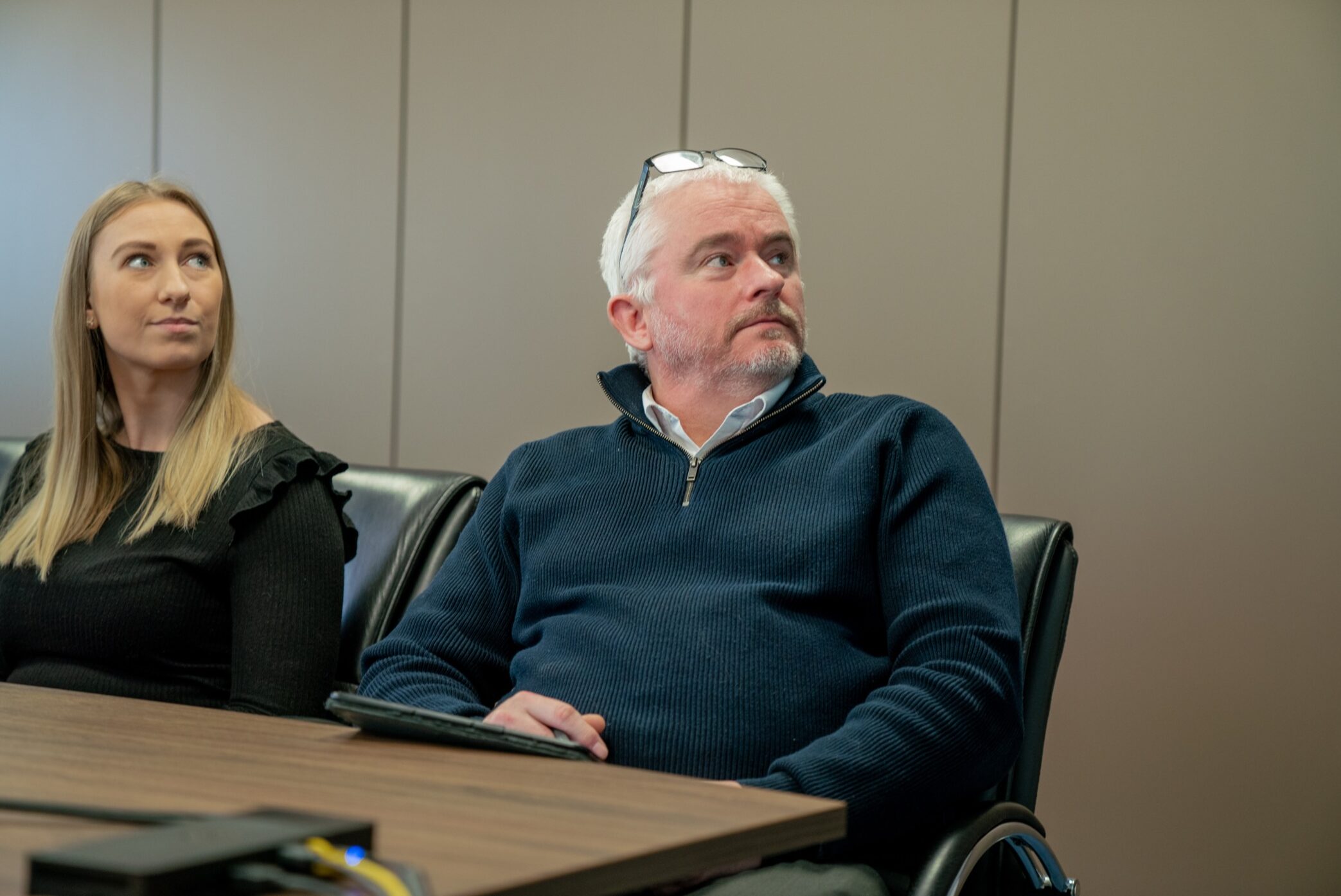 Enisca Browne - Two people in a meeting room sit at a table, attentively looking towards something off to the side.