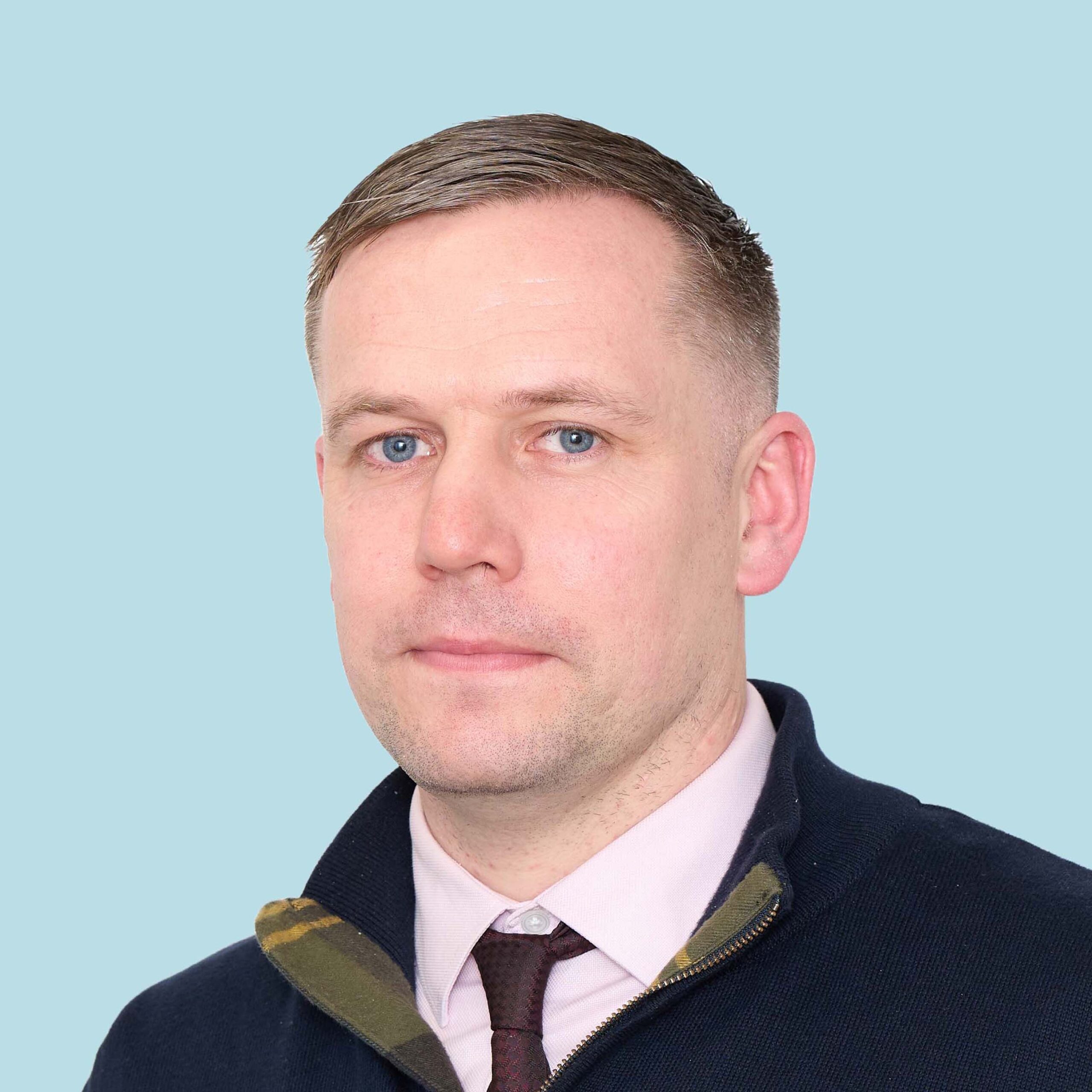 Enisca Browne - Man with short light brown hair and blue eyes wearing a navy jacket, pink shirt, and dark tie on light blue background.