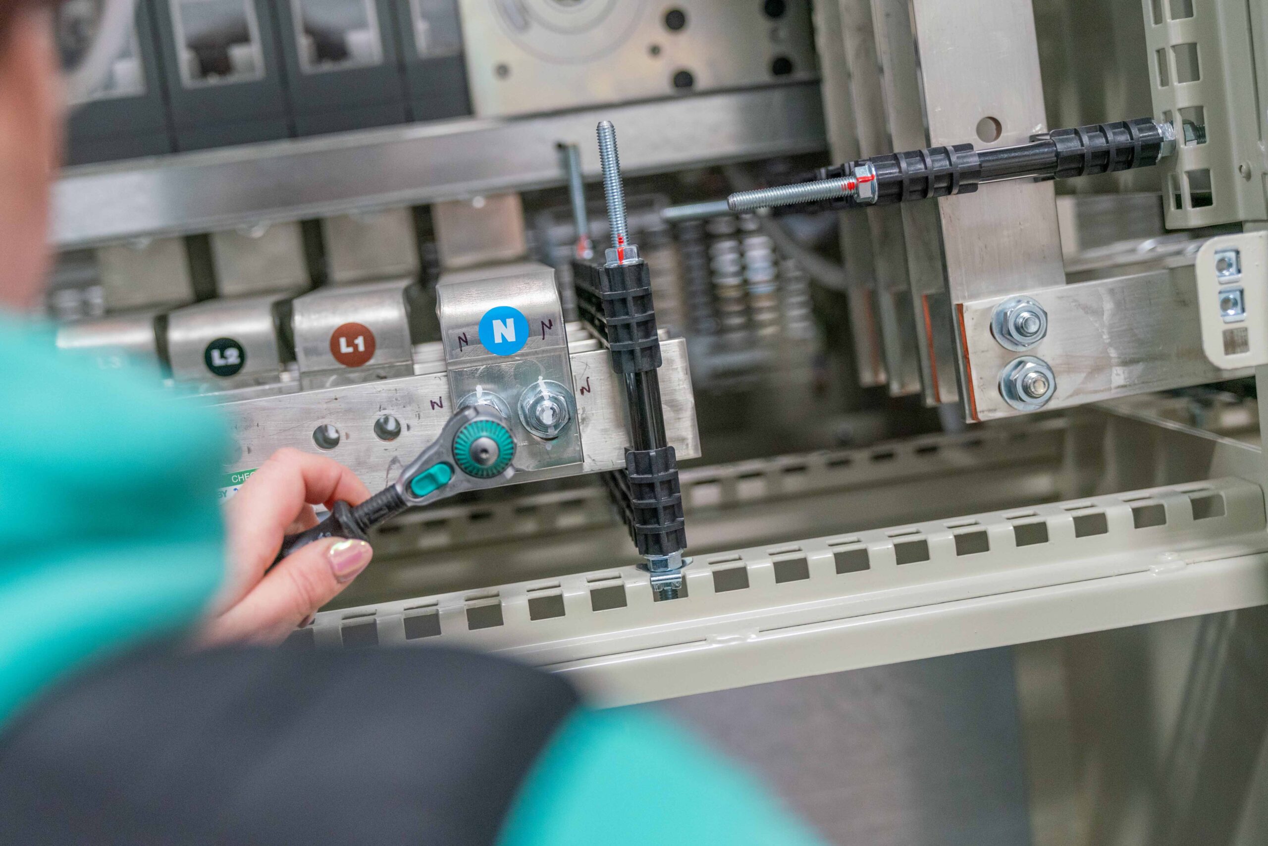 Enisca Browne - Close-up of a person using a wrench to adjust a bolt on an electrical panel with labeled terminals.