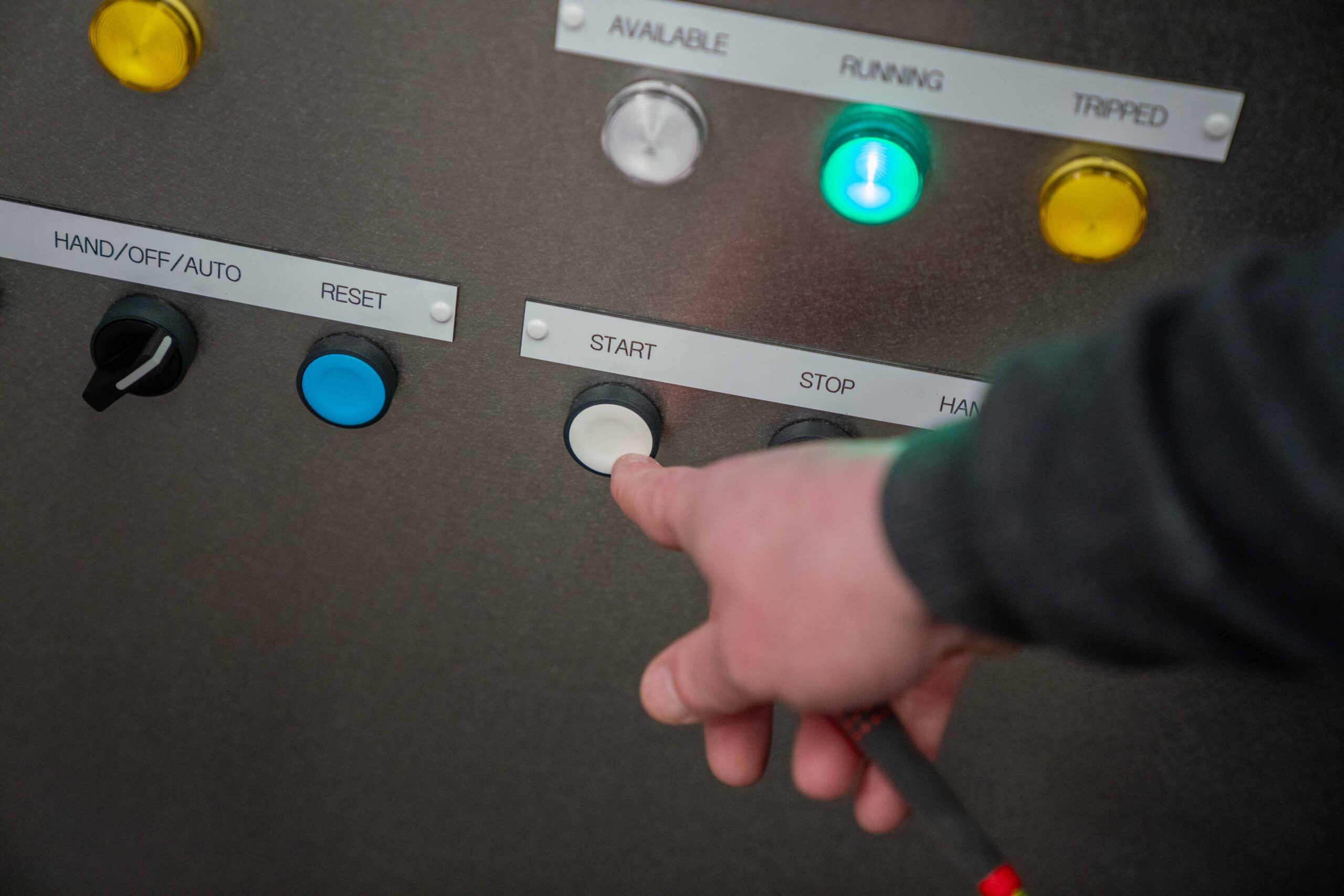Enisca Browne - A hand presses the white start button on an industrial control panel with labeled buttons and lights.