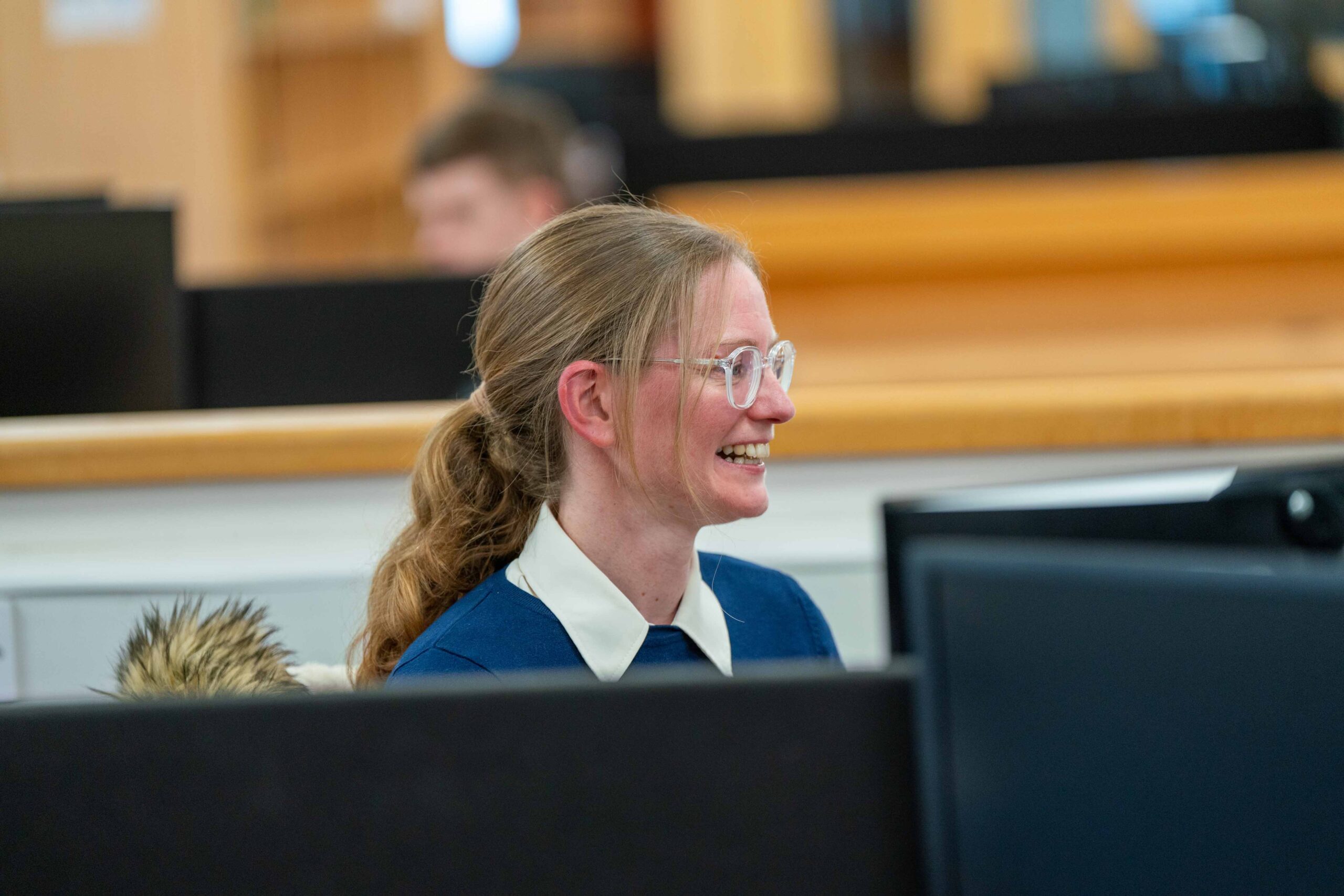 Enisca Browne - Smiling woman with glasses sits at a desk with computer screens in a bright office or library.