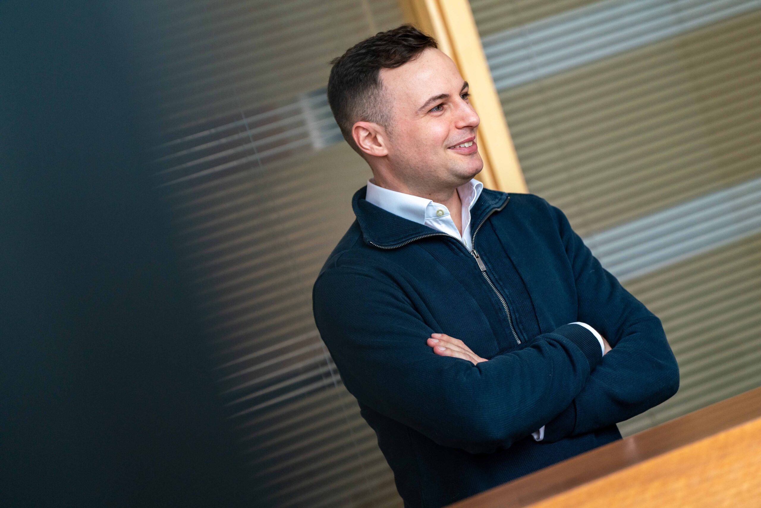 Enisca Browne - Man in a dark sweater stands with arms crossed, smiling, in an office setting with glass walls.