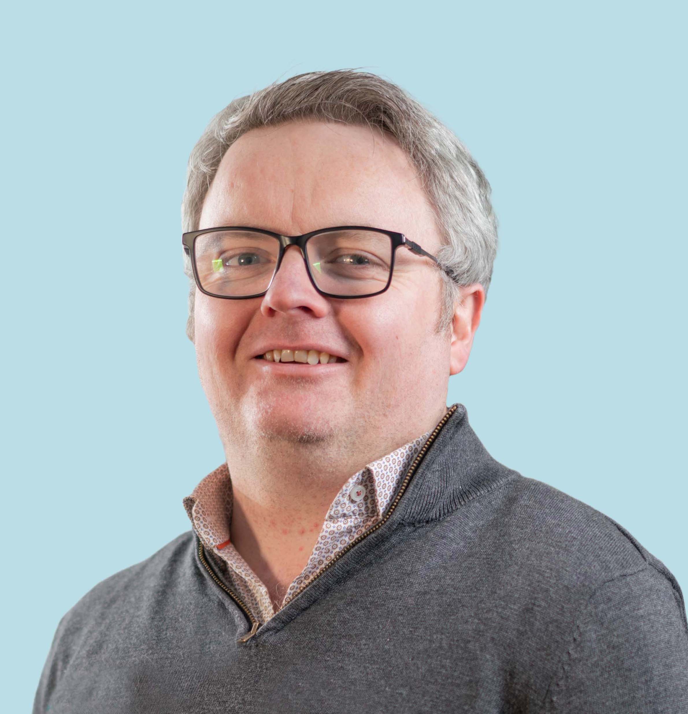 Enisca Browne - Smiling man with glasses and gray hair wearing a gray sweater, against a light blue background.
