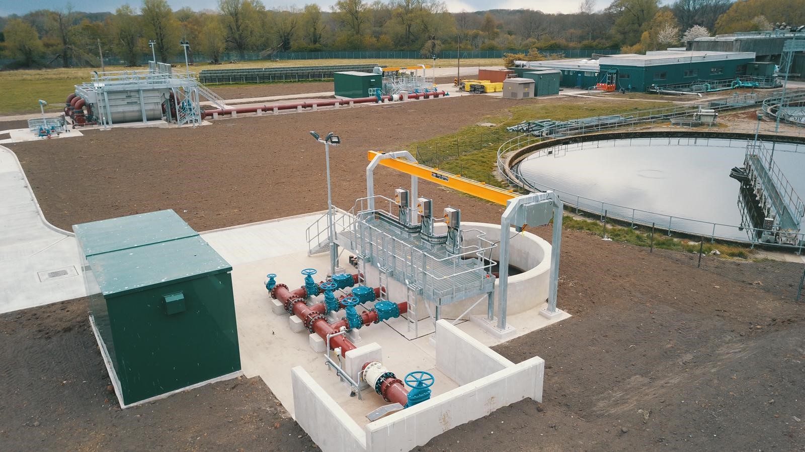 Enisca Browne - Aerial view of a water treatment facility with pipes, tanks, and green buildings surrounded by trees.