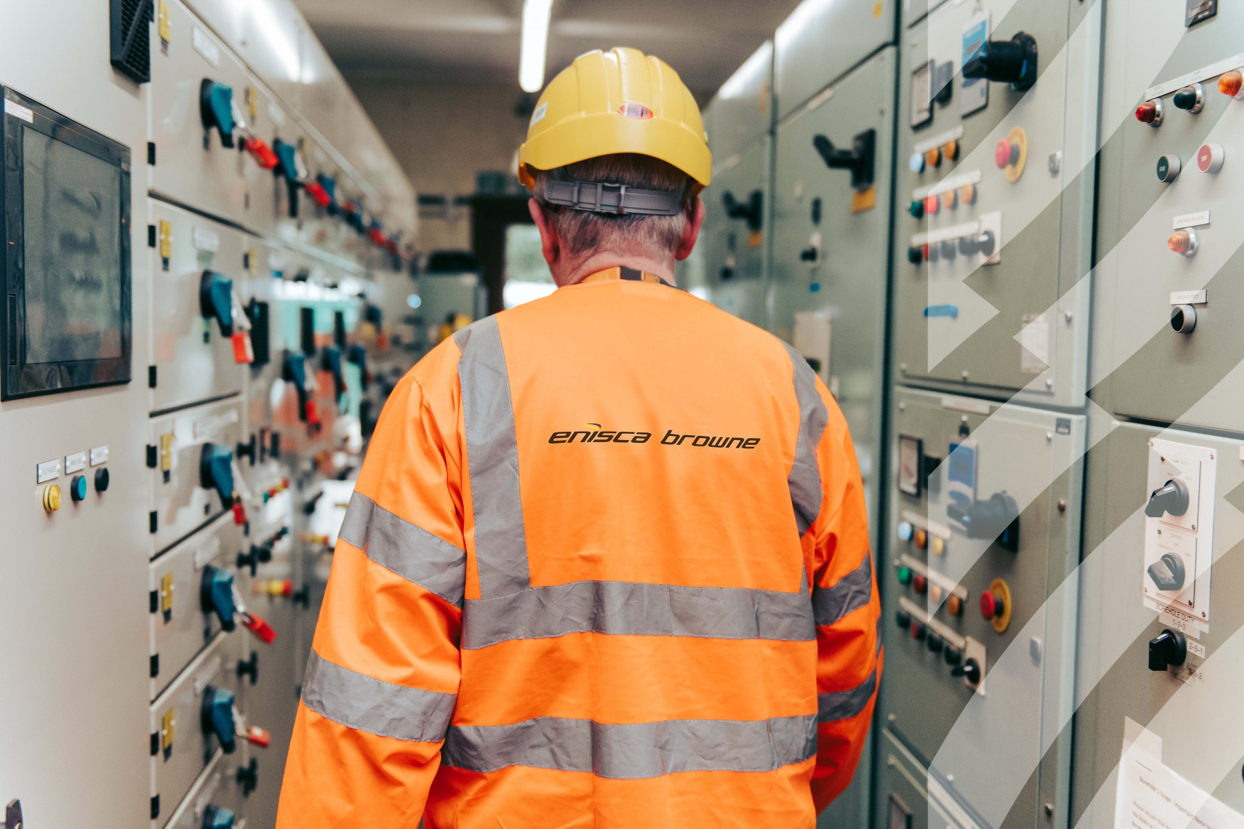 Enisca Browne - Worker in an orange safety jacket and helmet inspects control panels in an industrial facility.