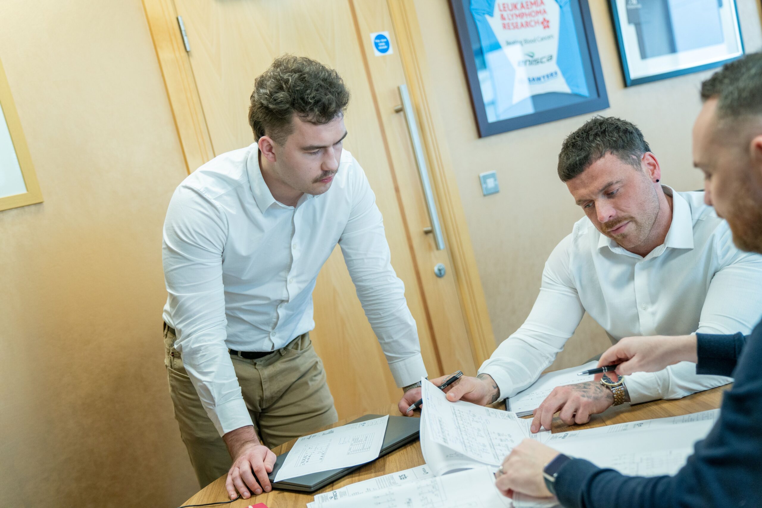 Enisca Browne - Three men in white shirts discuss documents and blueprints around a table in an office setting.