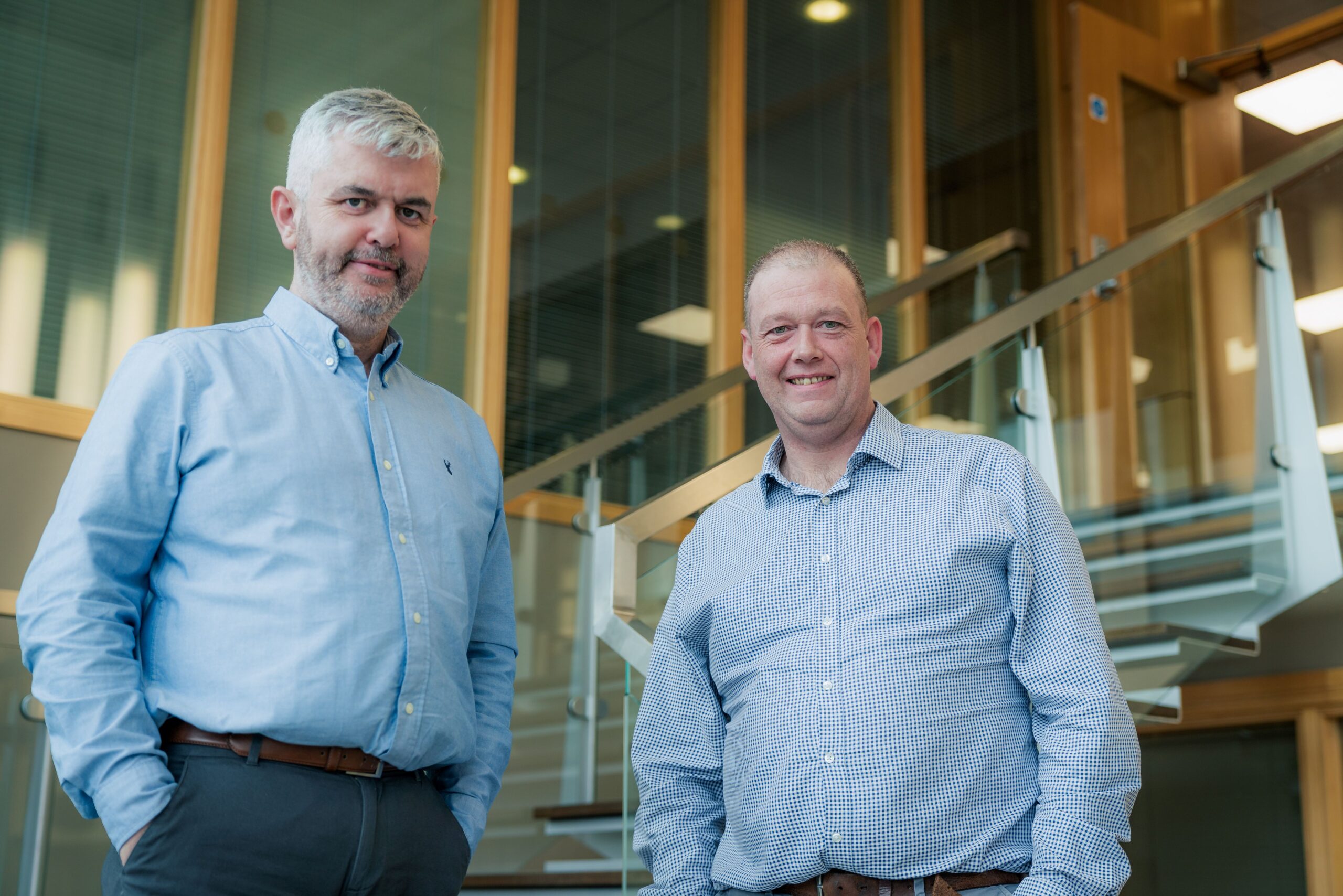 Enisca Browne - Two men in button-down shirts stand indoors in front of a modern staircase, smiling at the camera.