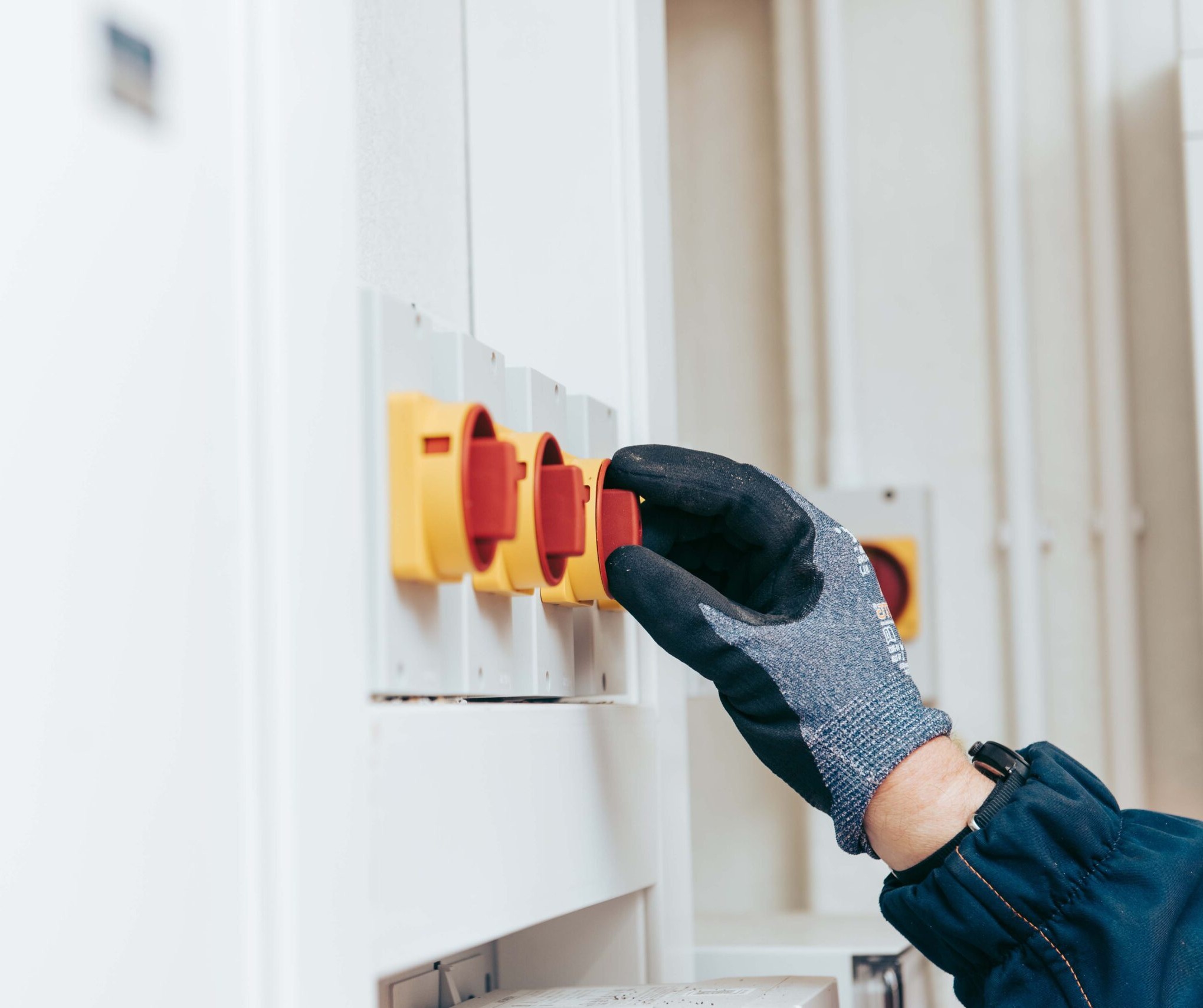 Enisca Browne - A gloved hand turns a red and yellow industrial switch on an electrical control panel.