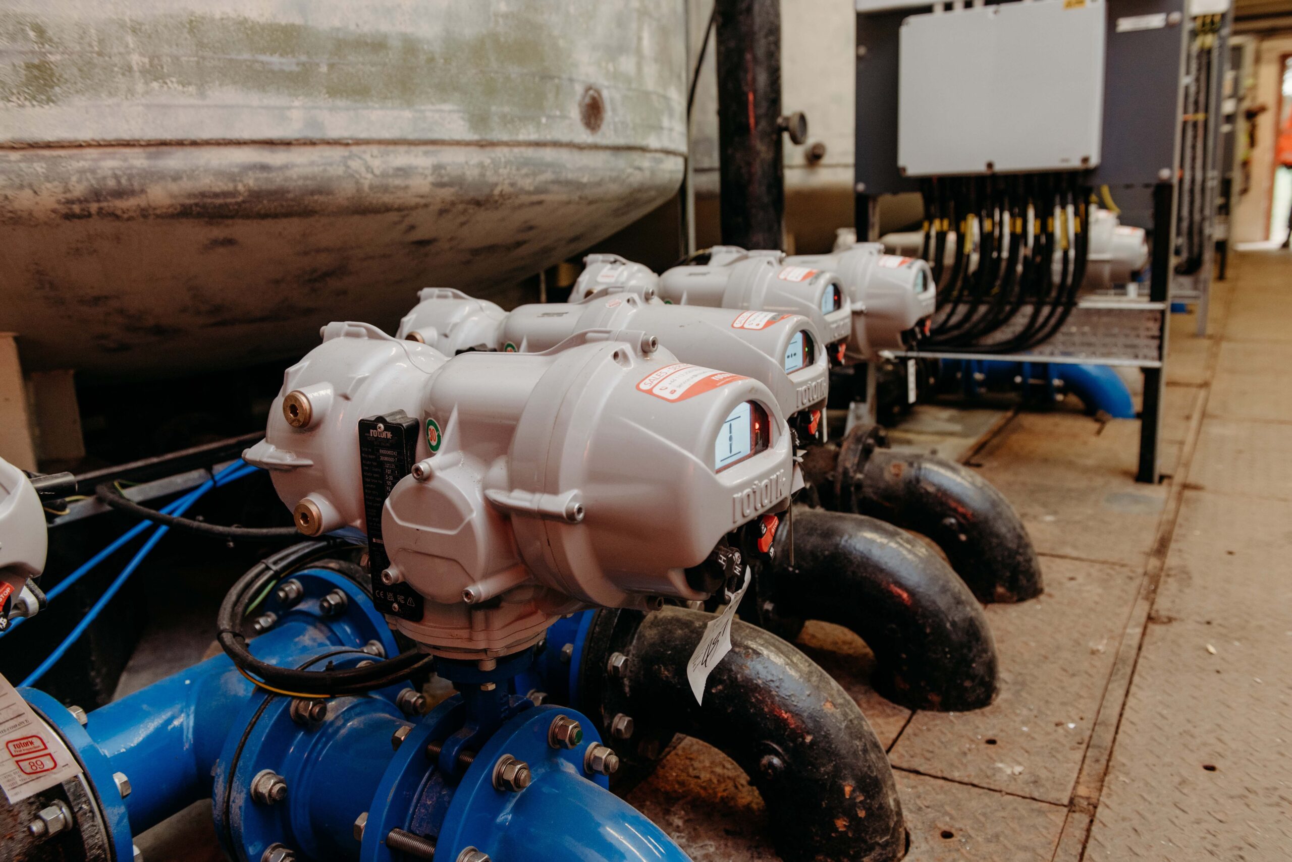 Enisca Browne - Rows of industrial valve actuators connected to blue pipes in a facility, with control panels nearby.