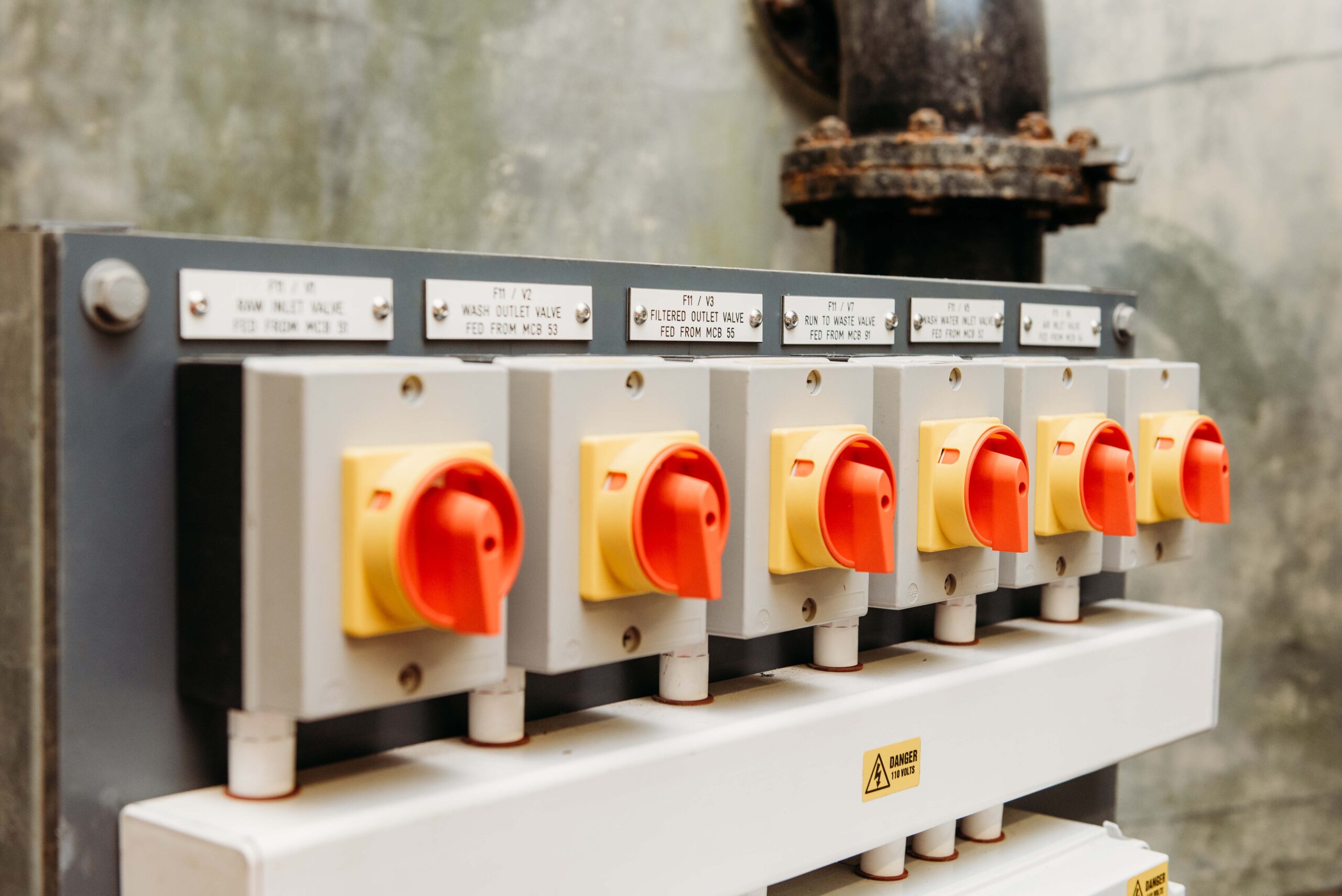 Enisca Browne - Six red and yellow industrial power switches mounted on a grey control panel against a concrete wall.