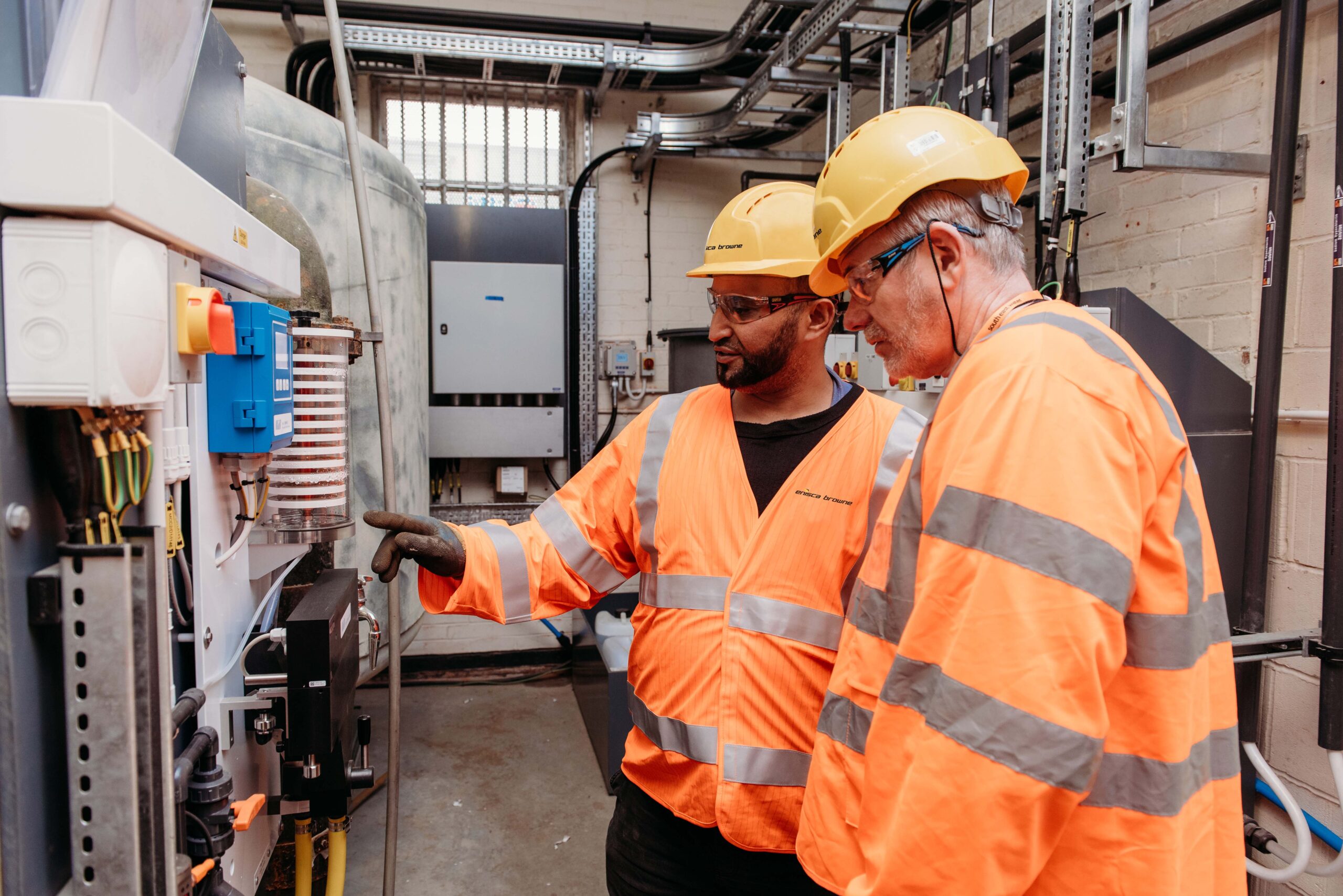 Enisca Browne - Two workers in orange safety gear and helmets inspect equipment in an industrial setting.