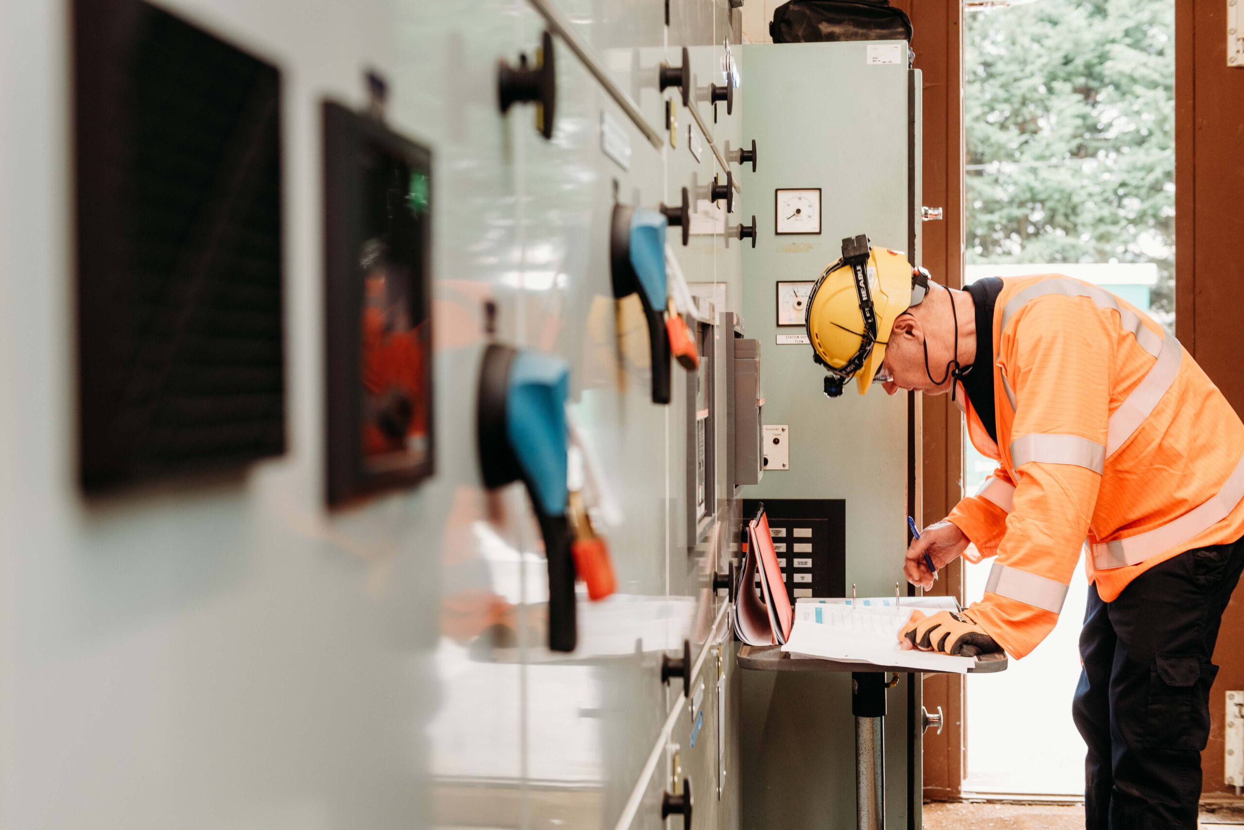 Enisca Browne - Engineer in a hard hat and hi-vis jacket inspects control panels and writes on a clipboard indoors.