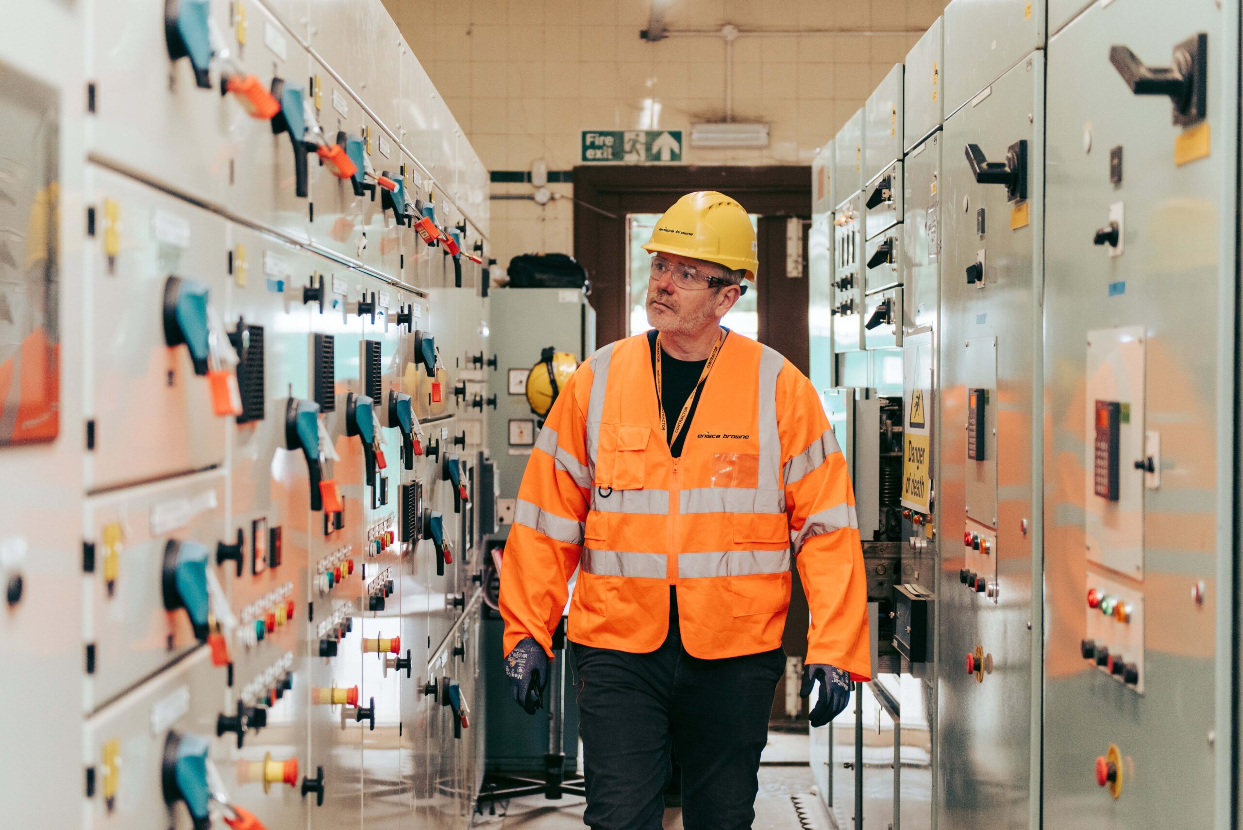 Enisca Browne - A worker in a hard hat and orange vest inspects control panels in an industrial facility.