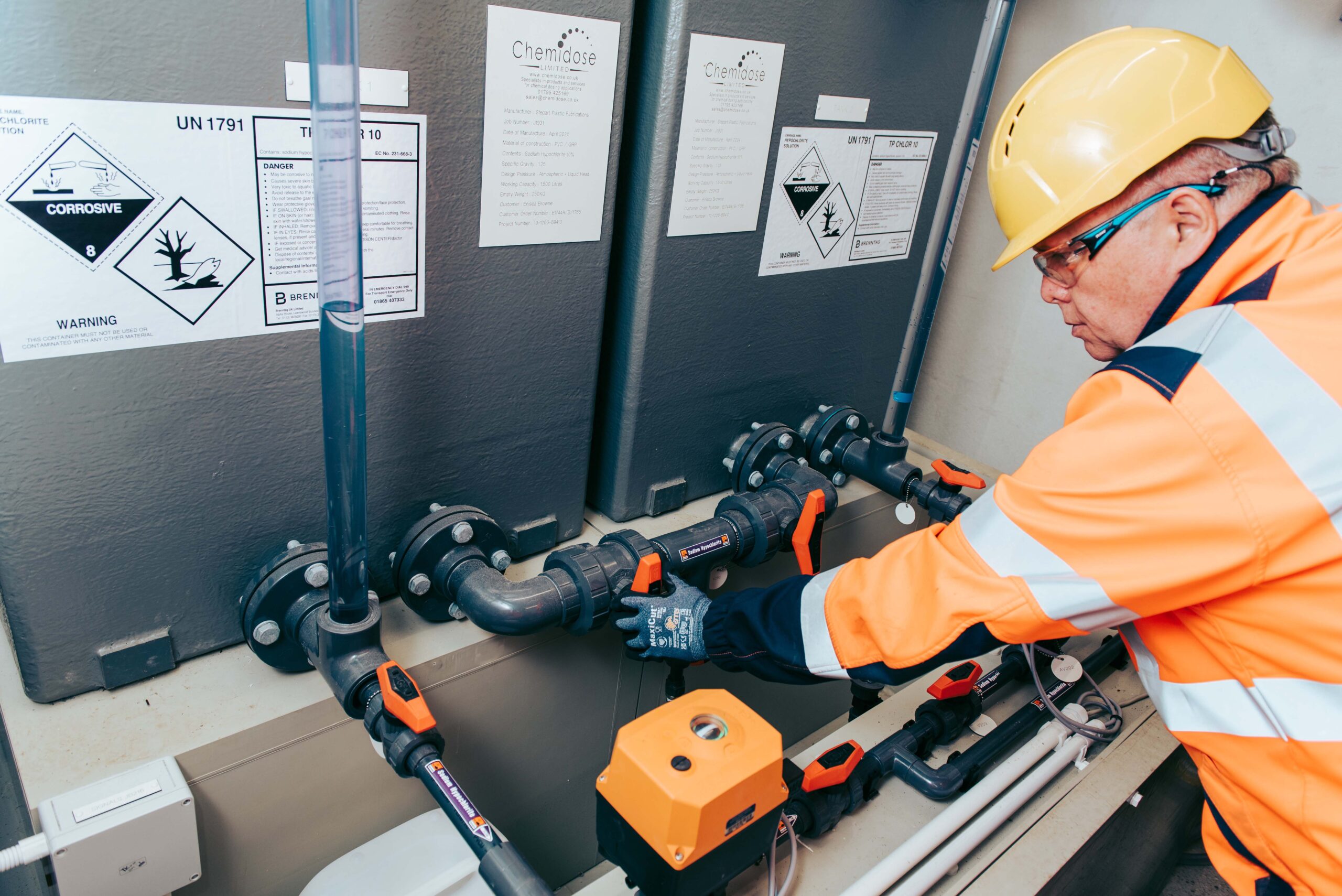 Enisca Browne - Worker in safety gear adjusts valves on industrial pipes near chemical warning signs.
