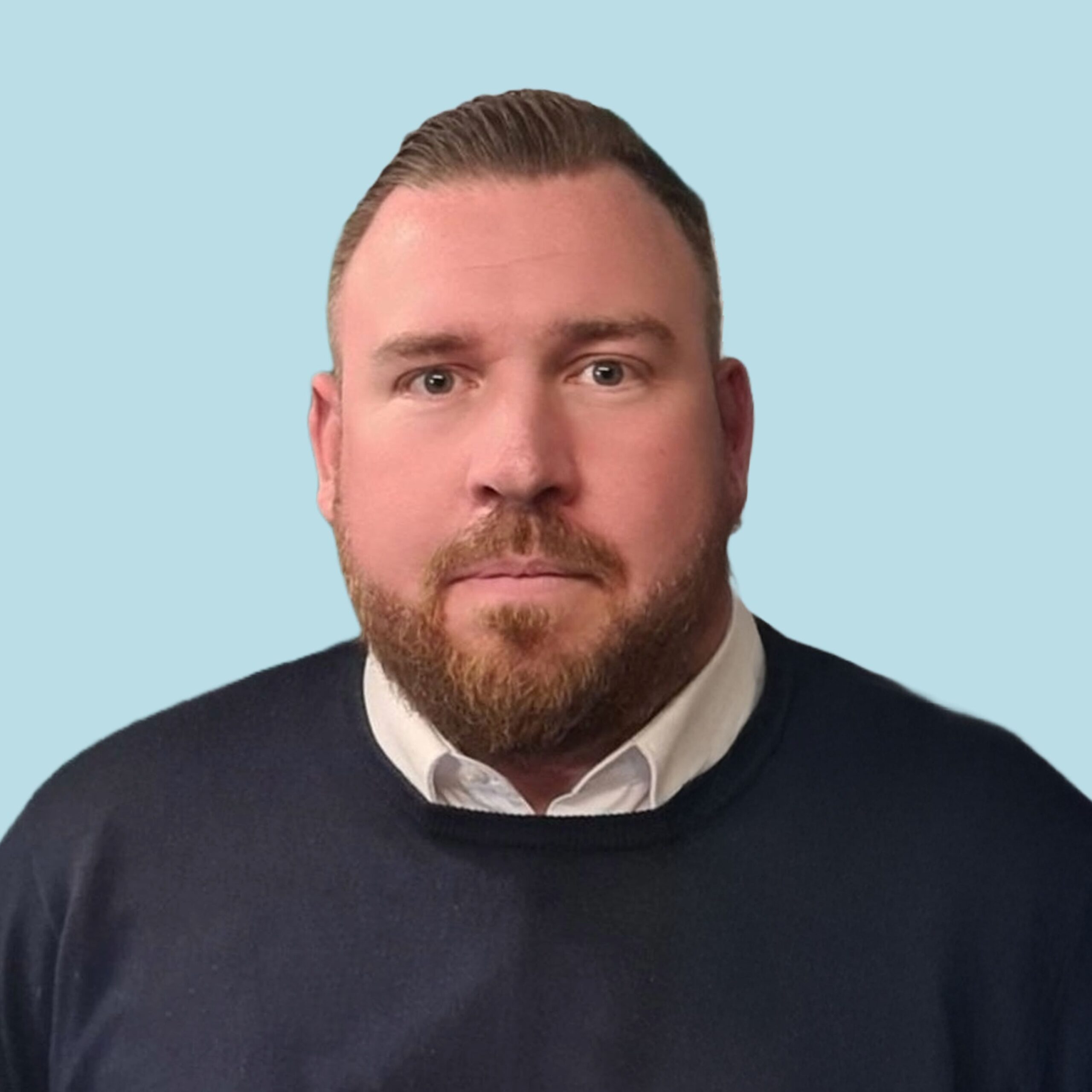 Enisca Browne - A man with short hair and a beard wearing a dark sweater and white shirt against a light blue background.
