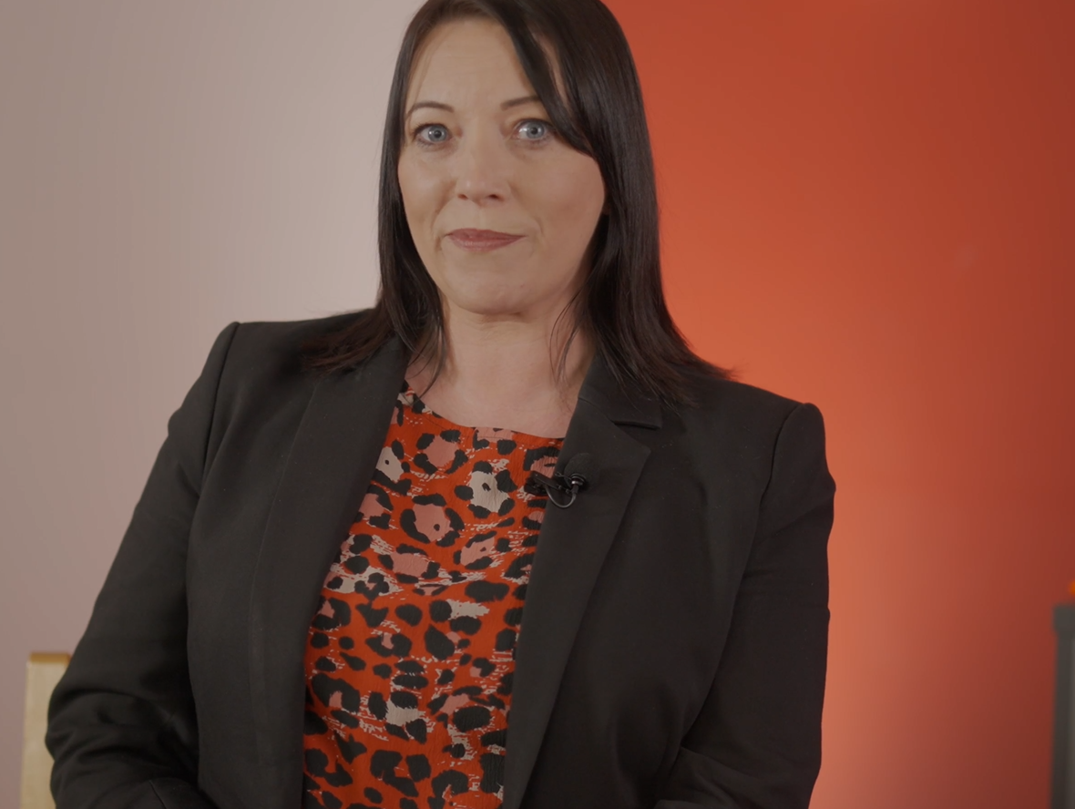 Enisca Browne - Woman with straight dark hair in a black blazer and red patterned top, sitting against an orange background.