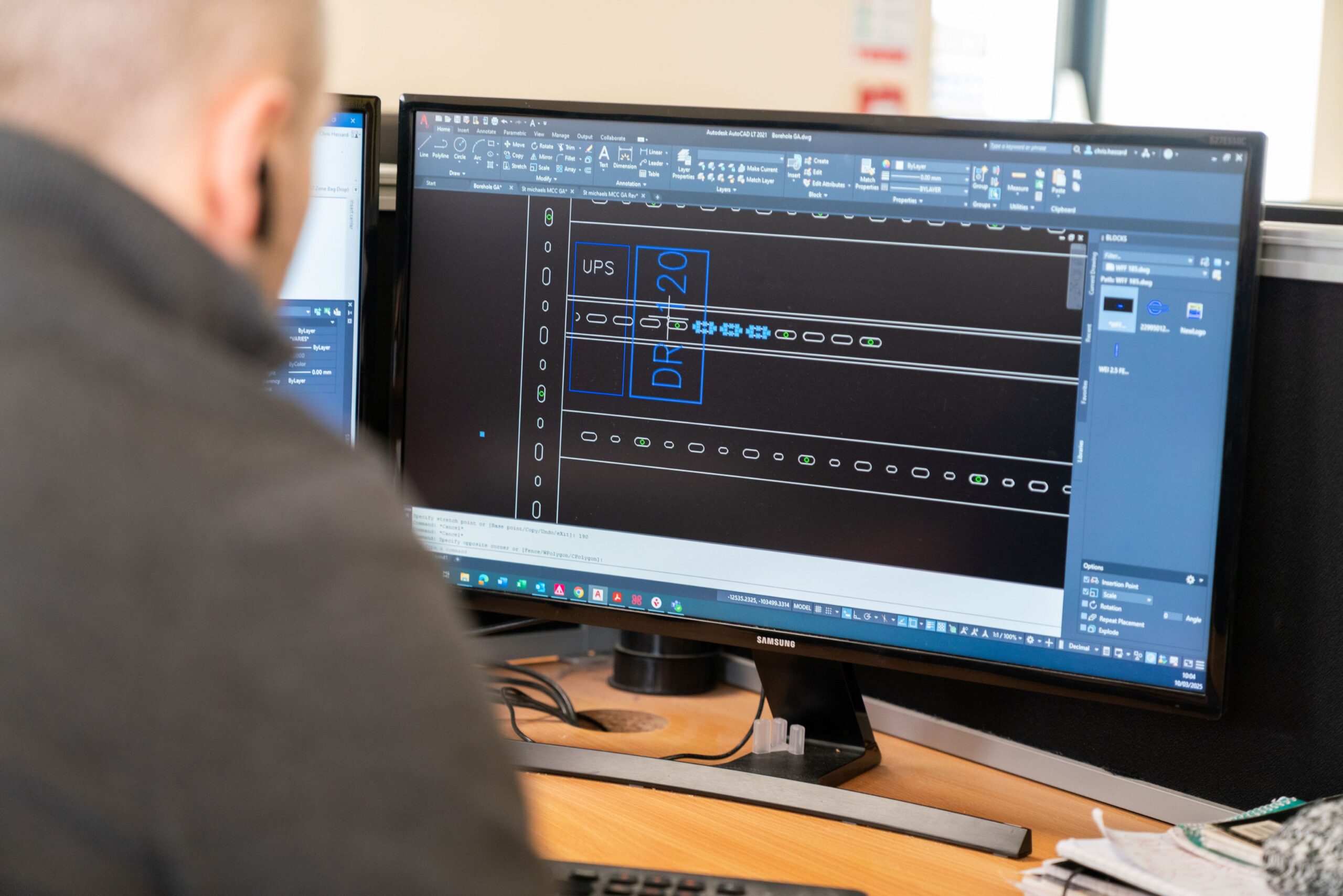 Enisca Browne - Person working at a desk with CAD software open on a large computer monitor displaying technical drawings.