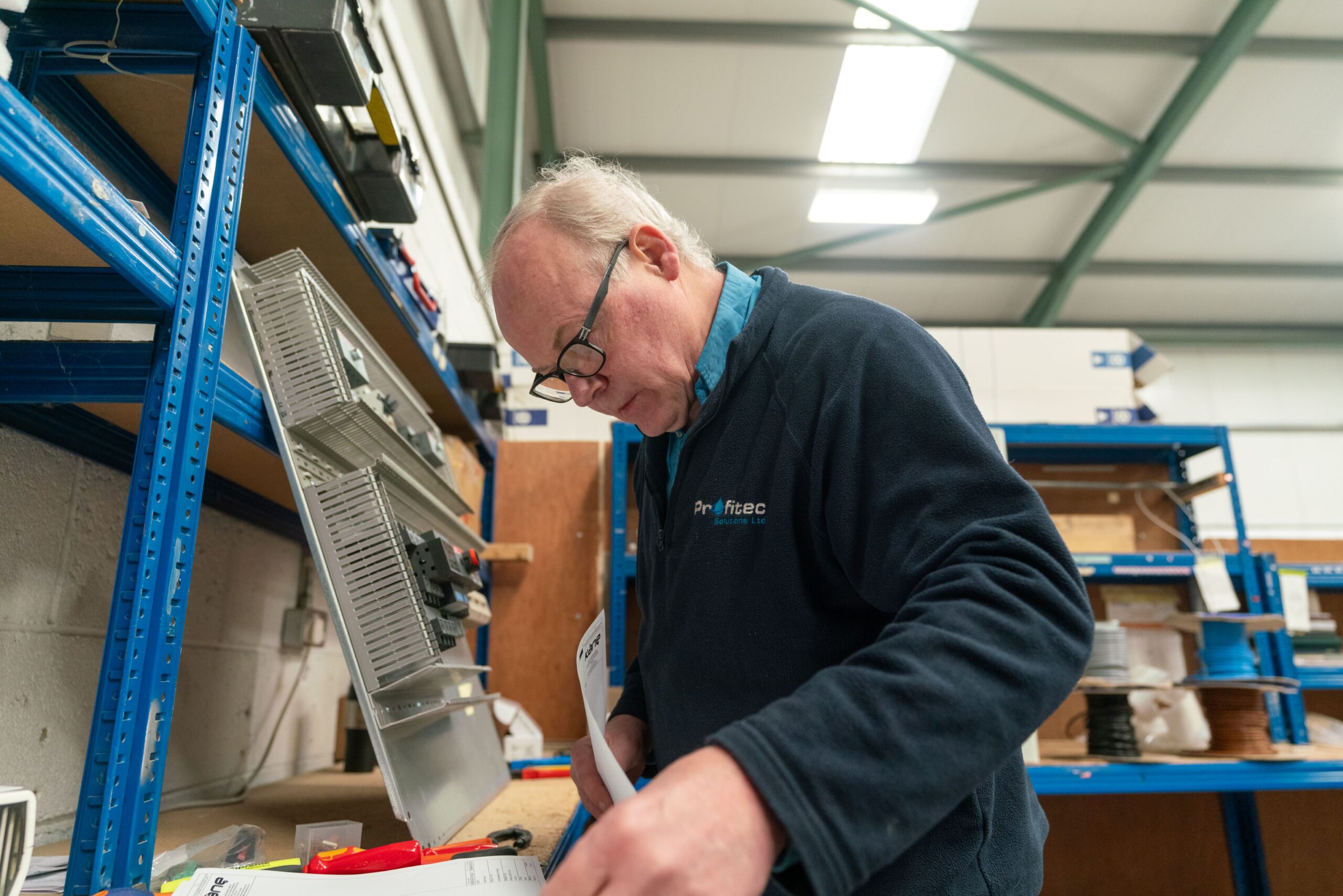 Enisca Browne - Older man with glasses works at a desk in a warehouse, examining equipment and paperwork.