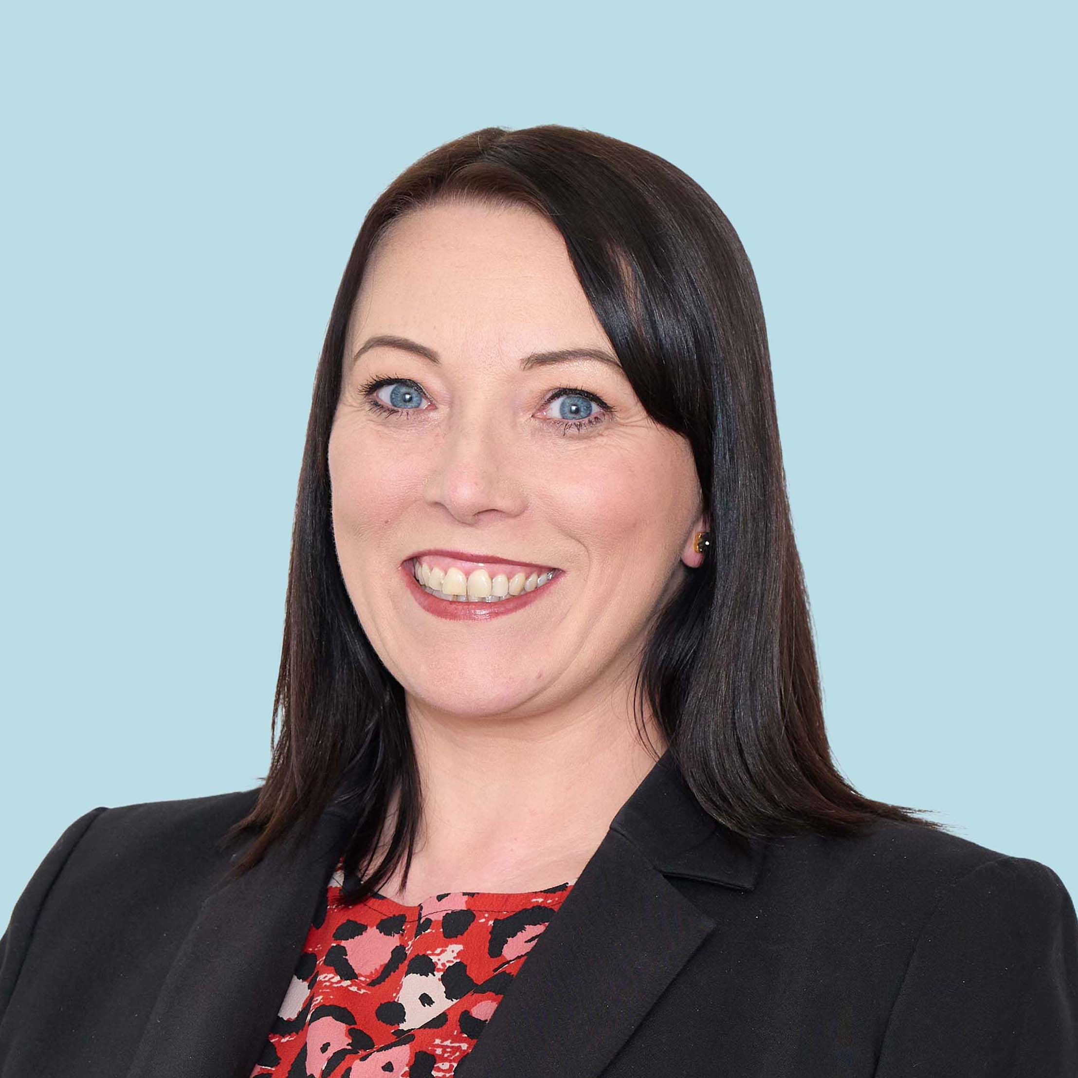 Enisca Browne - Smiling woman with straight dark hair wearing a black blazer and a red patterned top, on a light blue background.